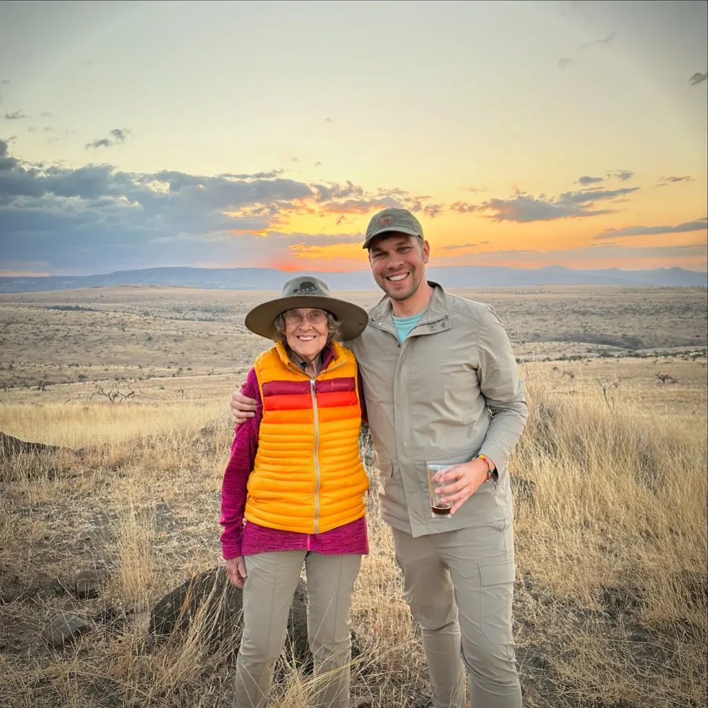 A man in a tan jacket and an older woman in a bright orange and yellow puffer vest stand together in a field of dry, golden grass. The woman wears a wide-brimmed safari hat. The background shows a vast, rolling landscape under a soft sunset sky.