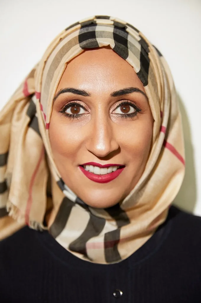 A professional portrait of Sam Latif, a woman with brown eyes and a friendly smile wearing a beige, black, and red checkered hijab. The headshot is a close-up against a plain white background, captured by photographer Holly McGlynn.
