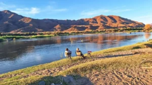 Two people sitting in camping chairs on a grassy riverbank, looking out over a wide, calm river. In the background, large, rugged mountains are bathed in the warm, golden light of late afternoon under a clear blue sky.