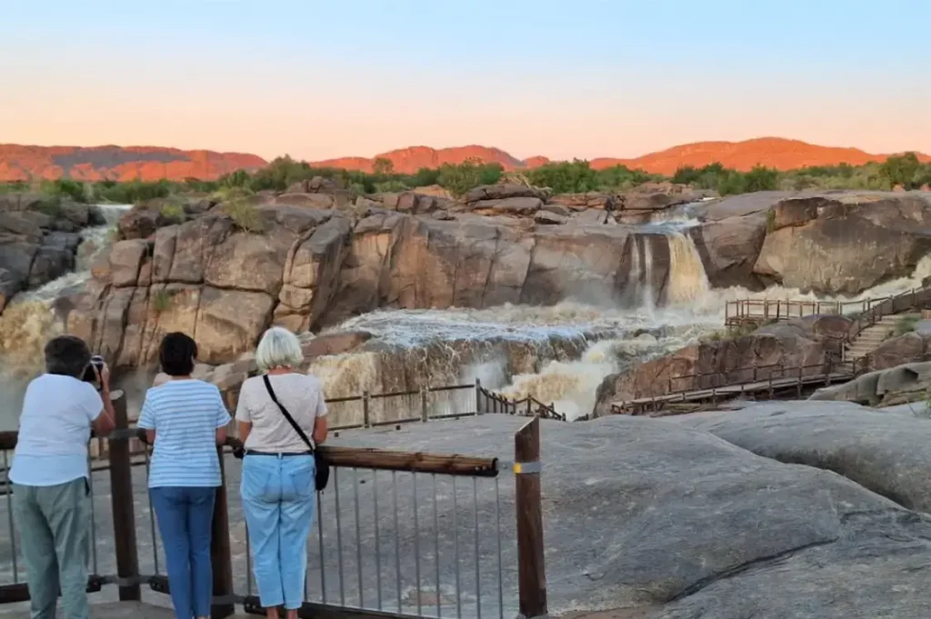 Two people sitting in camping chairs on a grassy riverbank, looking out over a wide, calm river. In the background, large, rugged mountains are bathed in the warm, golden light of late afternoon under a clear blue sky.