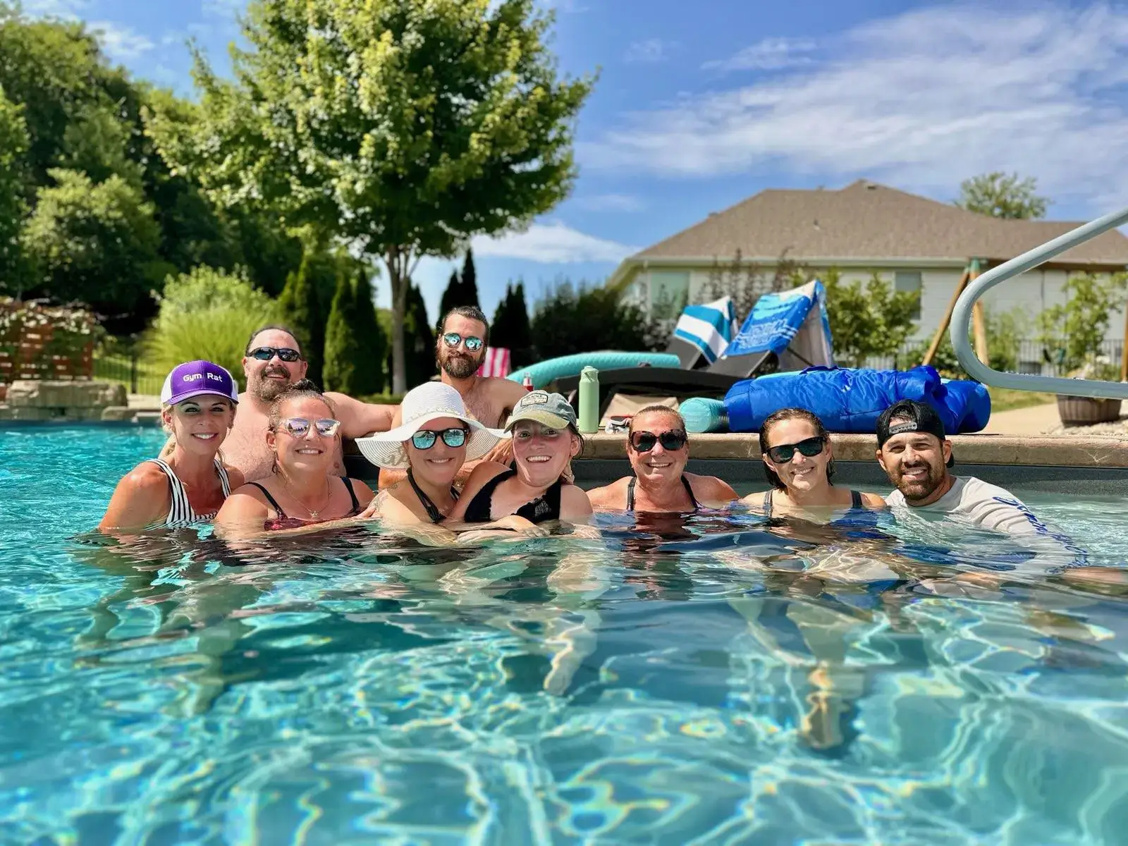 A group of eight friends wearing sunglasses and hats, smiling together while relaxing in a clear blue swimming pool on a sunny day in St. Louis.