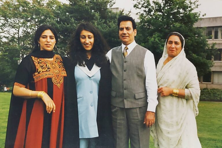 Four adults stand side by side outdoors on a grassy lawn, with trees and a building in the background. Two women on the left wear traditional-style dresses with gold detailing and layered fabrics, while the man beside them wears a grey vest over a white shirt and tie. The woman on the right wears a light-coloured outfit with a headscarf and gold jewelry. All face the camera with relaxed expressions.