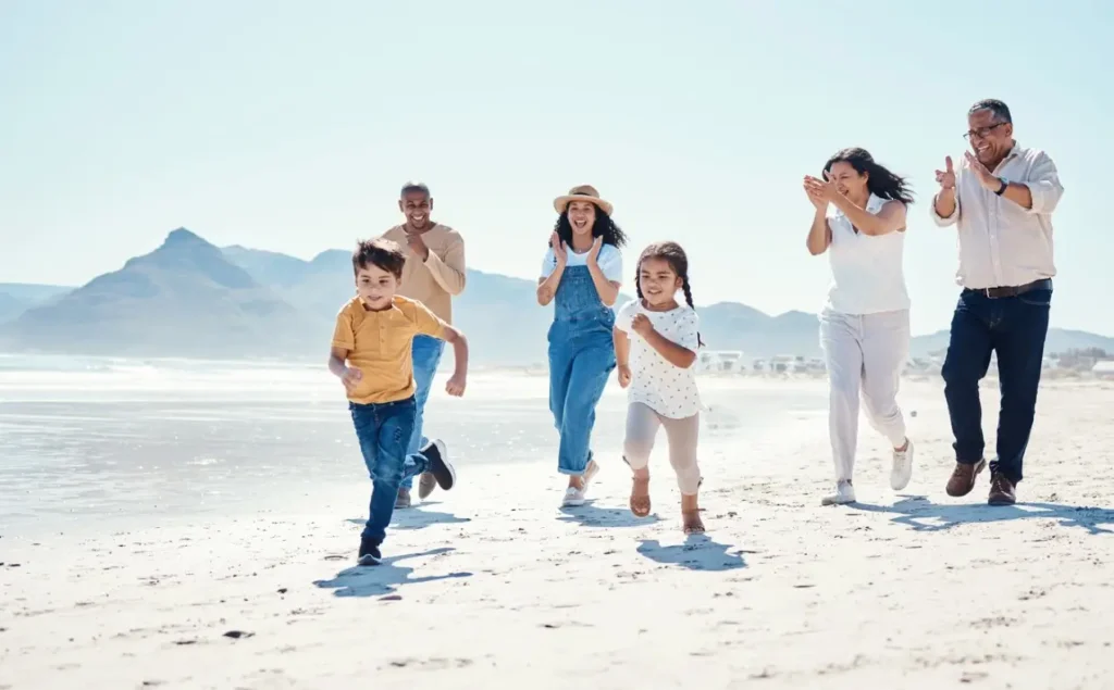 A multi-generational family happily running and playing on a sunny white sand beach with mountains in the background.