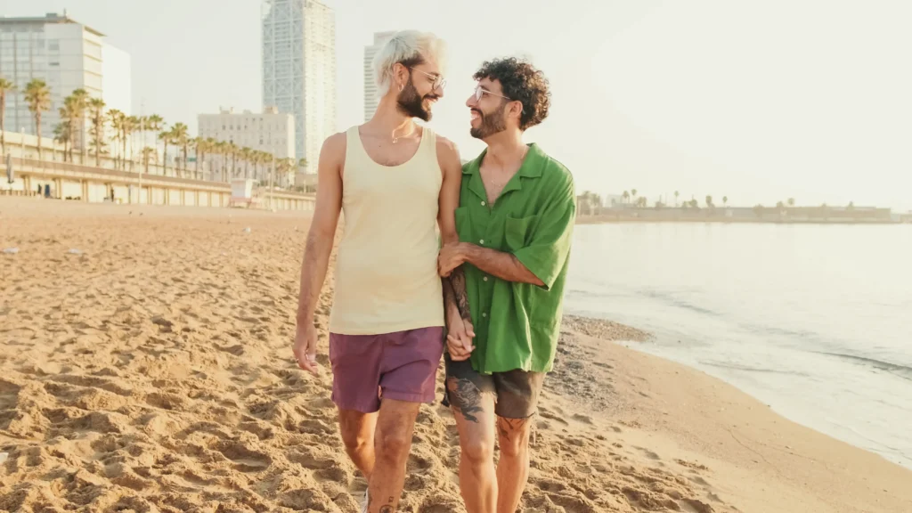 A happy gay couple walking hand-in-hand along a sandy beach at sunset, smiling at each other with a city skyline and palm trees in the background.