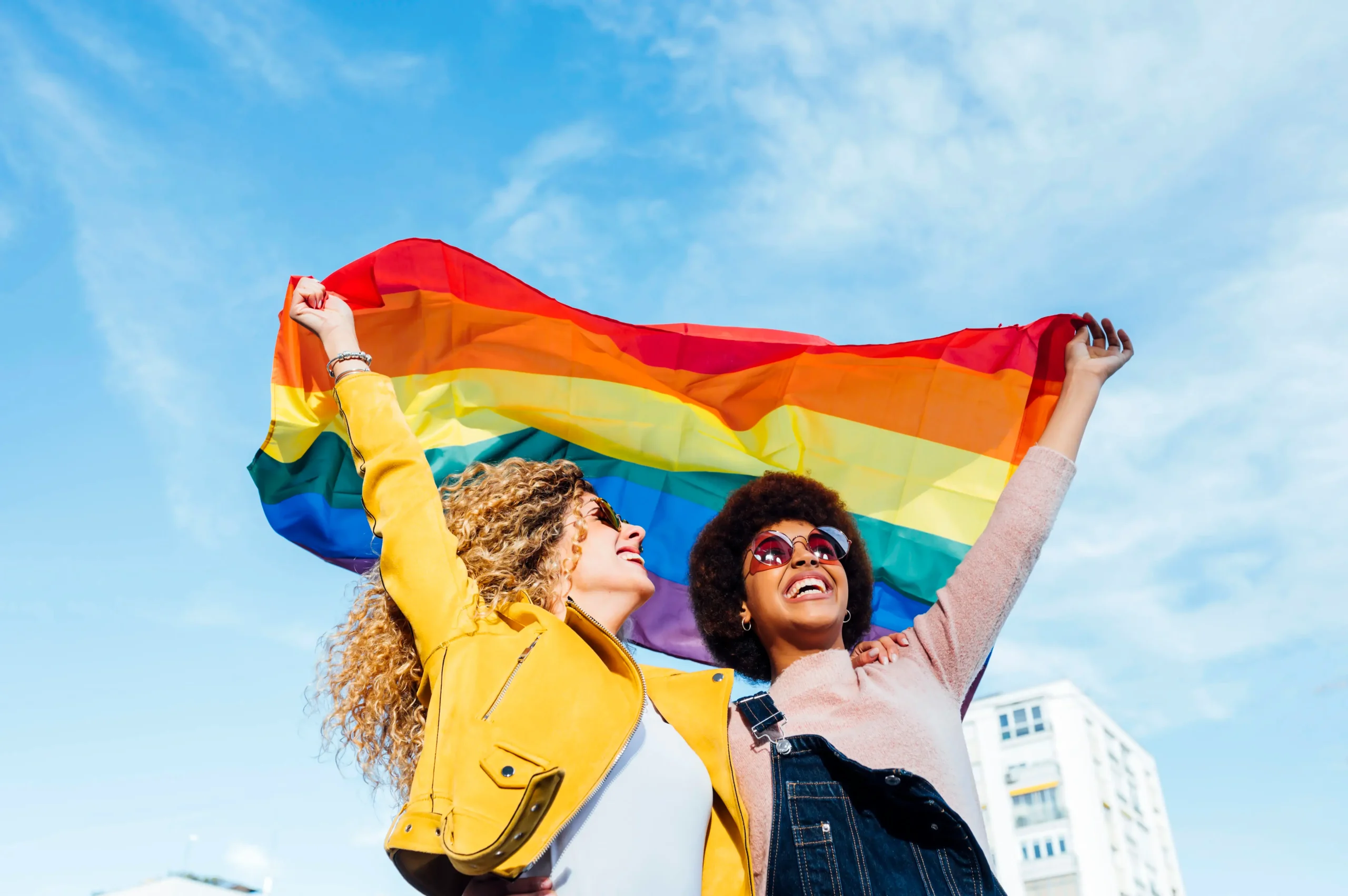 Two joyful women celebrating outdoors, holding a large rainbow Pride flag aloft against a clear blue sky and city buildings.