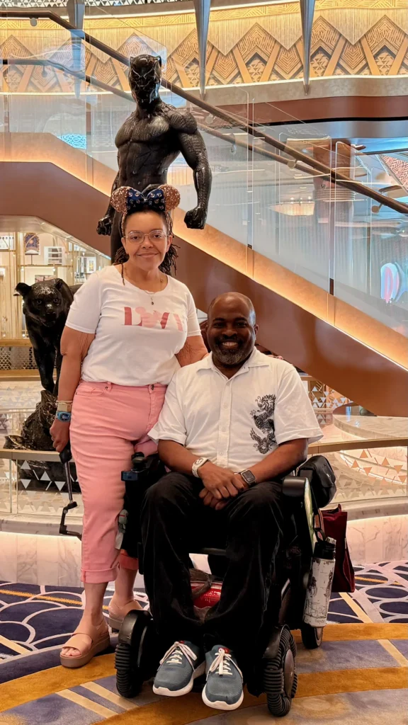 Aadam, a smiling Black man using a power wheelchair, and Brooke, wearing pink pants and sparkly Minnie Mouse ears, posing together next to a large bronze Black Panther statue on a Disney Cruise Line ship.