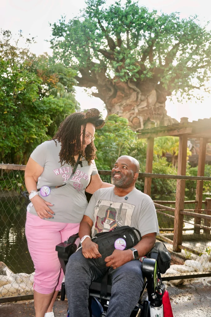Brooke and her husband Aadam, a wheelchair user, sharing a warm look in front of the Tree of Life at Disney's Animal Kingdom. Brooke wears leopard-print Minnie Mouse ears, and both wear Disney celebration buttons.