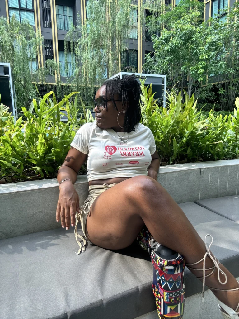 A Black woman wearing sunglasses and a white t-shirt sitting on an outdoor lounge bench in front of lush green plants, showing her colorful patterned prosthetic leg.