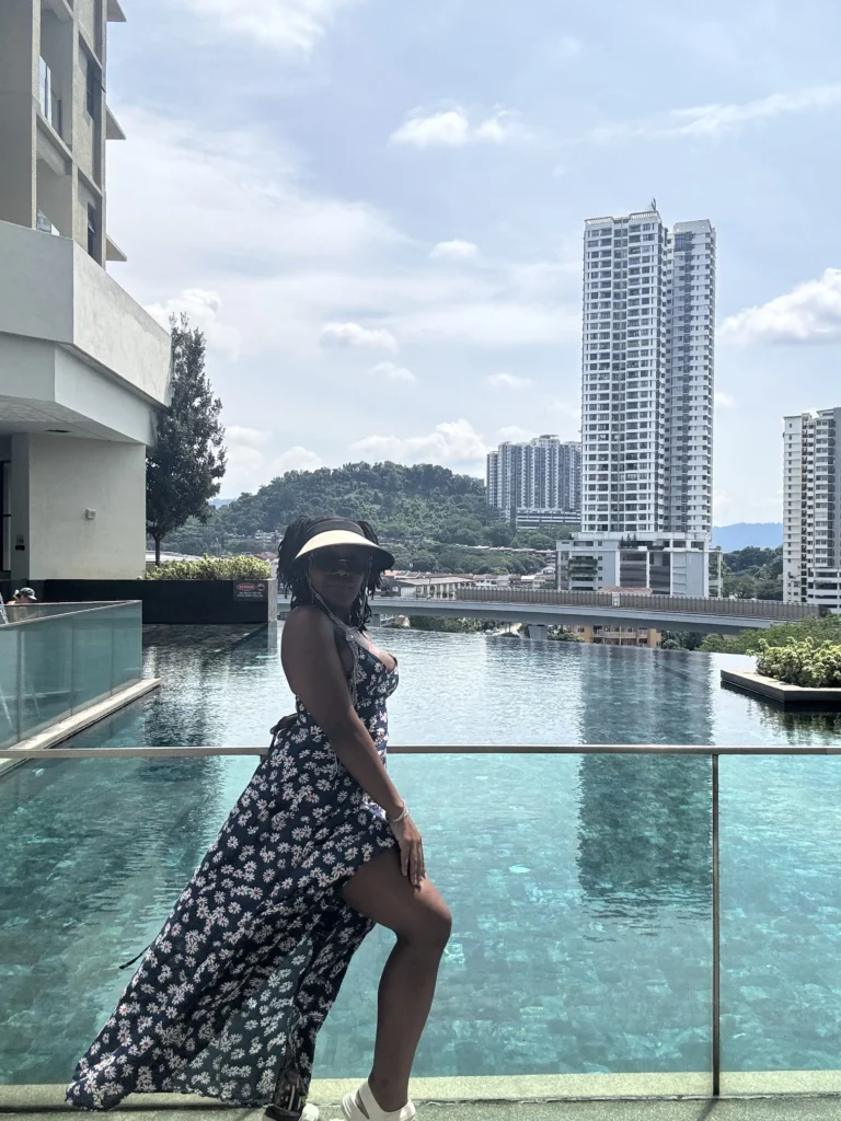 A Black woman wearing a floral maxi dress and sun visor posing by a rooftop infinity pool overlooking city skyscrapers and green hills.