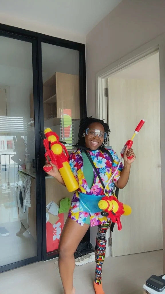 A happy Black woman with a colorful prosthetic leg wearing a floral shirt and holding bright yellow water guns during a Songkran water festival celebration.
