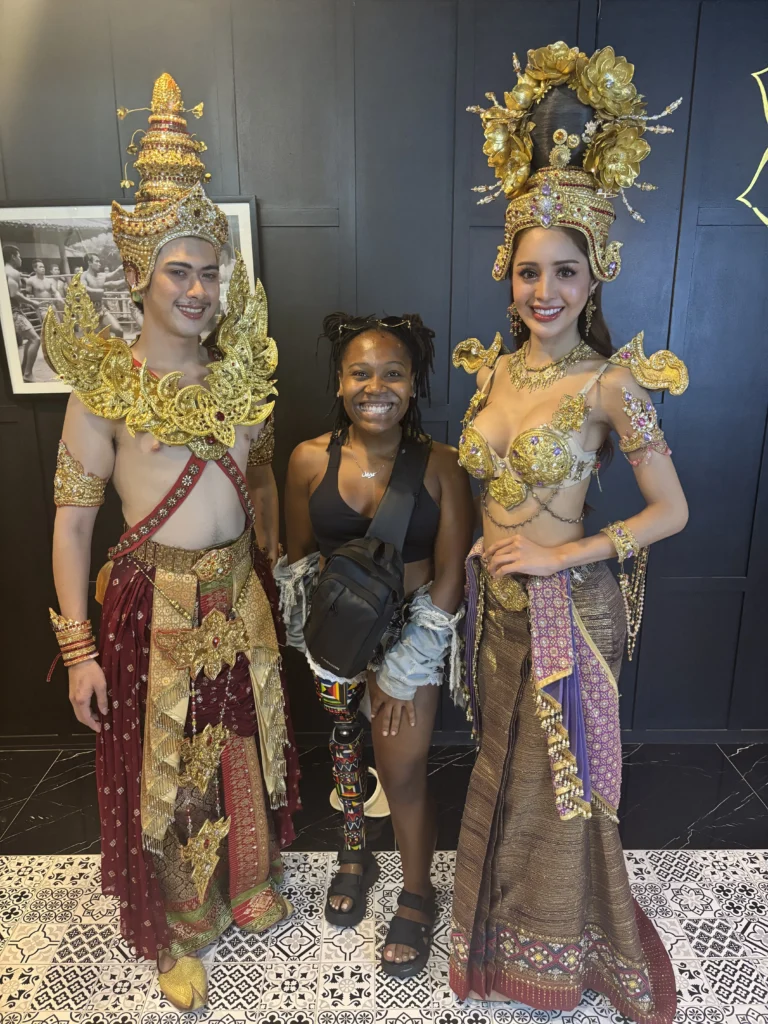 A smiling Black woman with a prosthetic leg standing between two performers wearing elaborate, traditional golden Thai costumes with ornate headdresses.