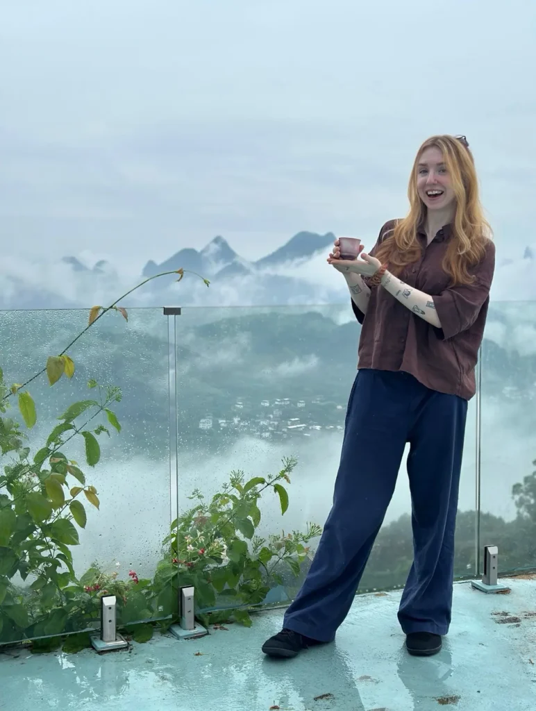 Solo traveler Amy Aed smiles, holding a local tea cup against a dramatic, foggy mountain landscape in rural China.
