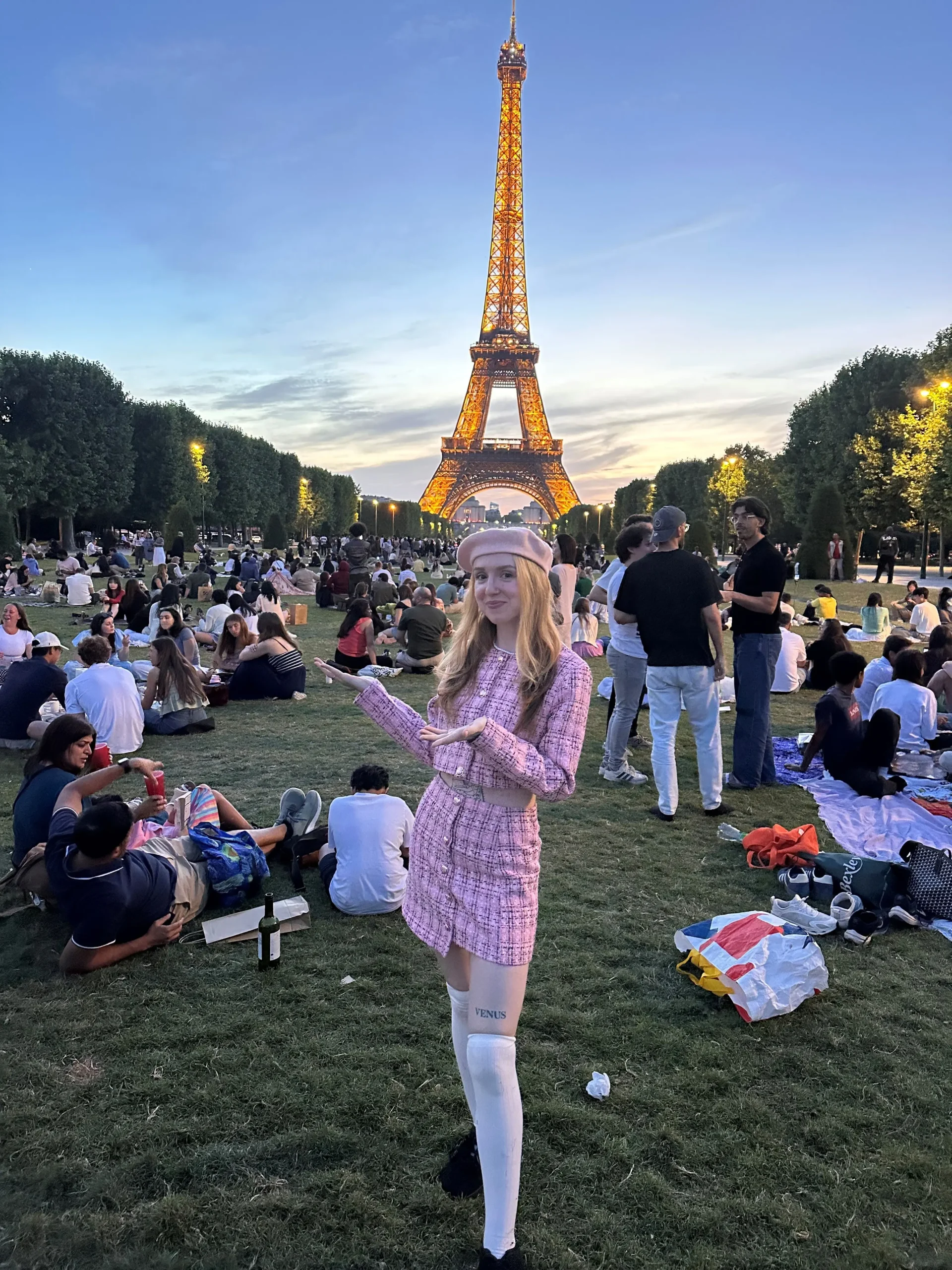 Travel blogger Amy Aed in a chic pink tweed outfit and beret, gesturing towards the illuminated Eiffel Tower at sunset.