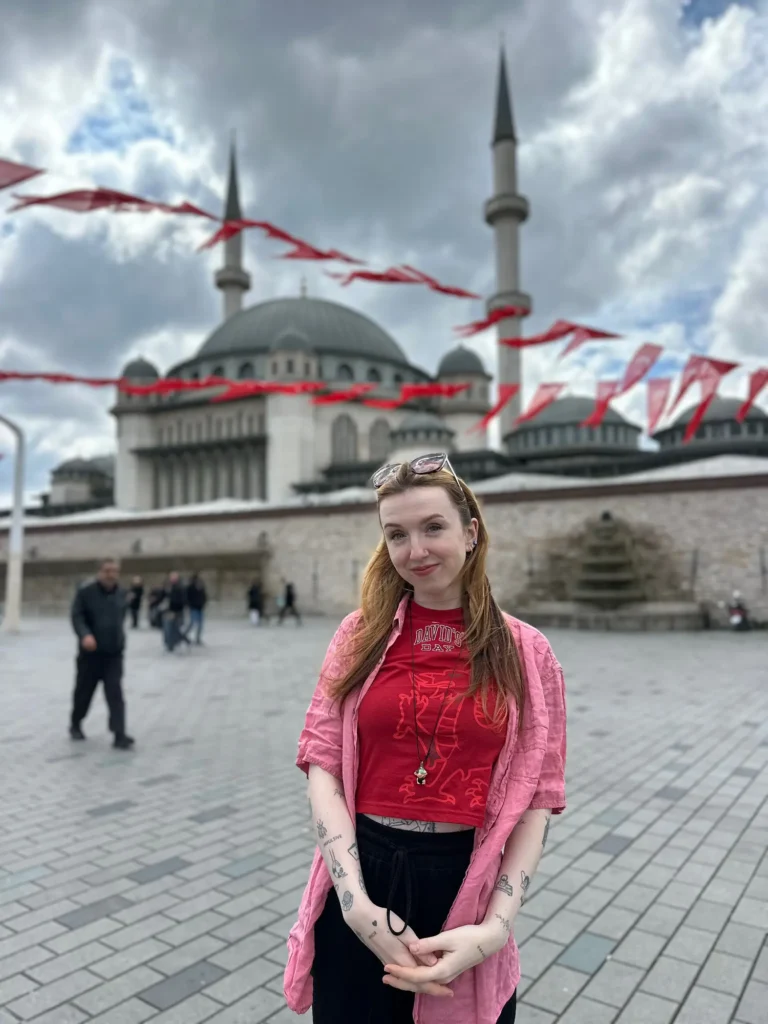 Amy Aed, smiling and wearing a pink overshirt, stands in front of the grand Taksim Mosque in Istanbul, Turkey.