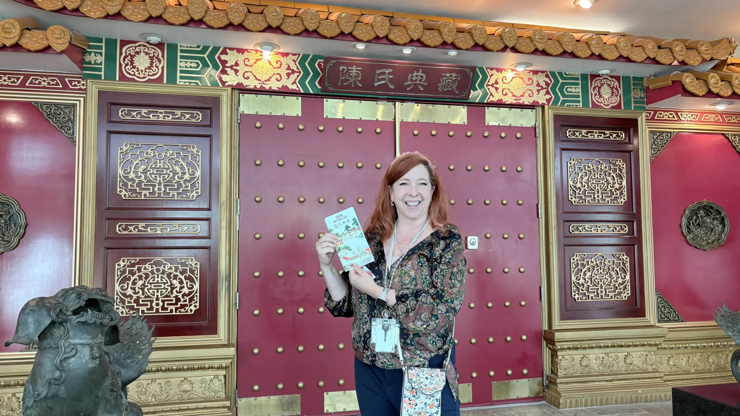 A woman holding a gallery brochure smiling in front of ornate, traditional red and gold Chinese entrance doors at the Chen Art Gallery in Torrance, California.