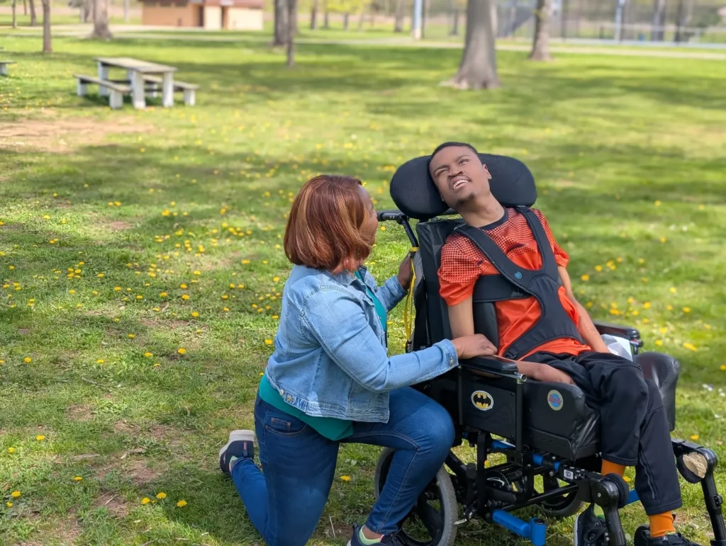 Christine Staple Ebanks kneeling on a grassy lawn in a park, smiling warmheartedly while looking at a young man resting in an supportive outdoor wheelchair.