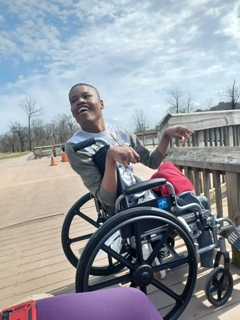 A smiling young Black man seated in a manual wheelchair outdoors on an accessible wooden boardwalk under a partly cloudy sky.