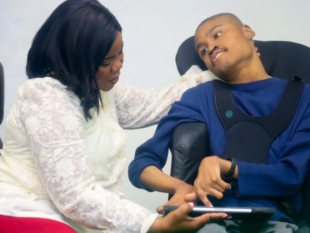 Christine Staple Ebanks kneeling beside a young Black man in a specialized supportive wheelchair as they interact with a digital tablet together indoors.