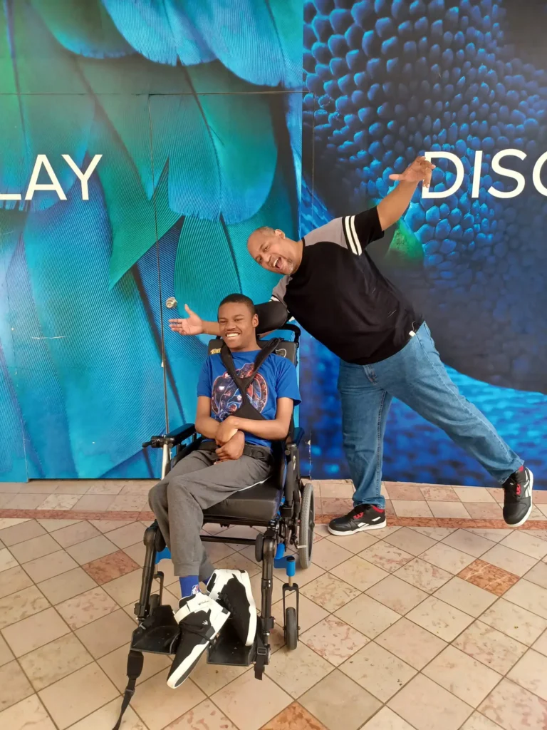 A happy young Black man in a supportive wheelchair smiling alongside a playful adult male family member posing enthusiastically in front of a blue feather pattern wall decor.