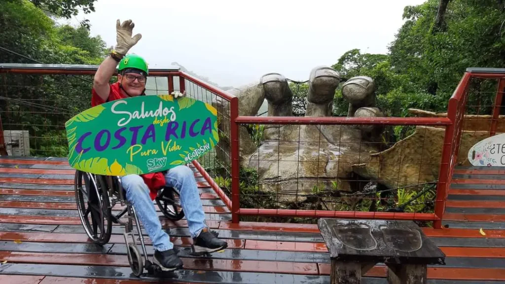 A smiling man in a wheelchair wearing a safety helmet and waving next to a large stone hand sculpture at an accessible viewing platform in Costa Rica.