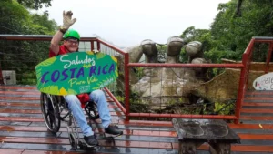 A smiling man in a wheelchair wearing a safety helmet and waving next to a large stone hand sculpture at an accessible viewing platform in Costa Rica.