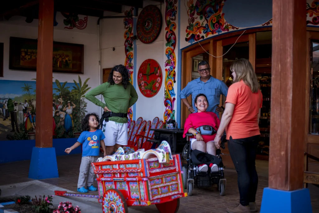 A smiling family, including a woman in a wheelchair, visiting a colorful, hand-painted traditional oxcart display at an accessible tourist attraction.