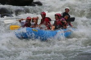 A group of excited tourists wearing red helmets and life jackets paddling a blue inflatable raft through white water rapids.