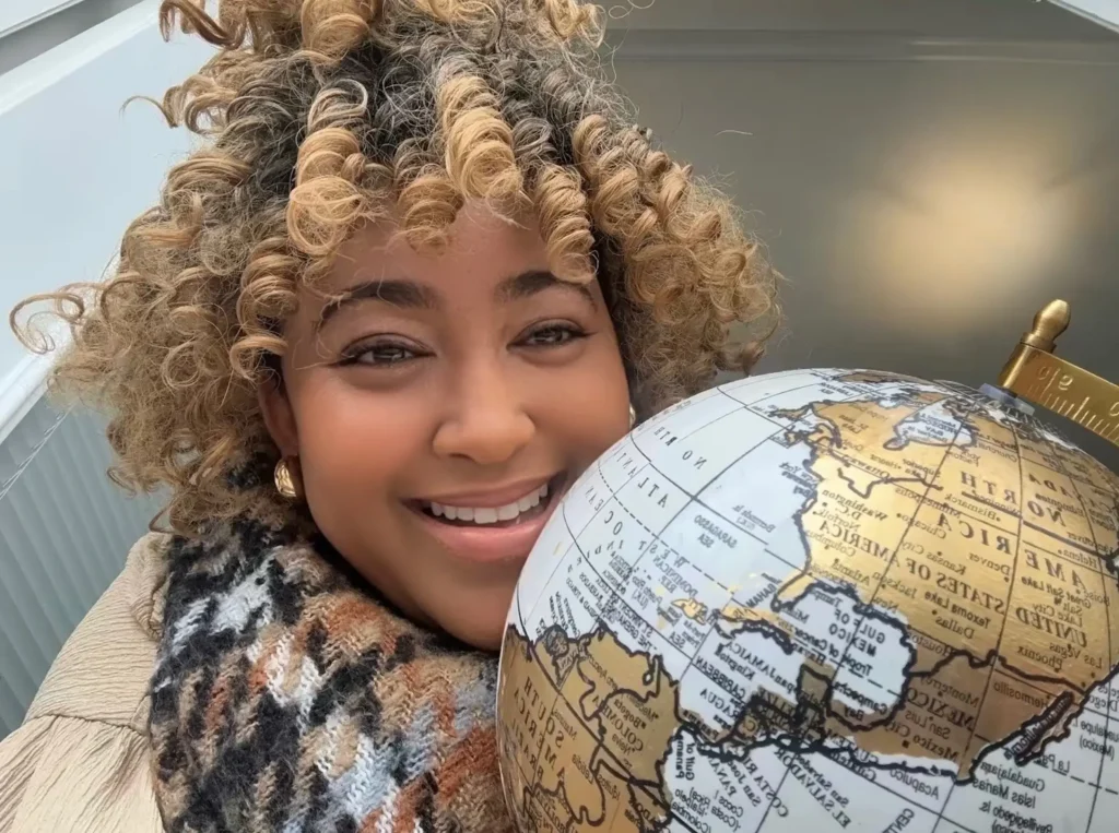 Smiling woman with blonde curly hair holding a vintage gold desktop globe indoors.