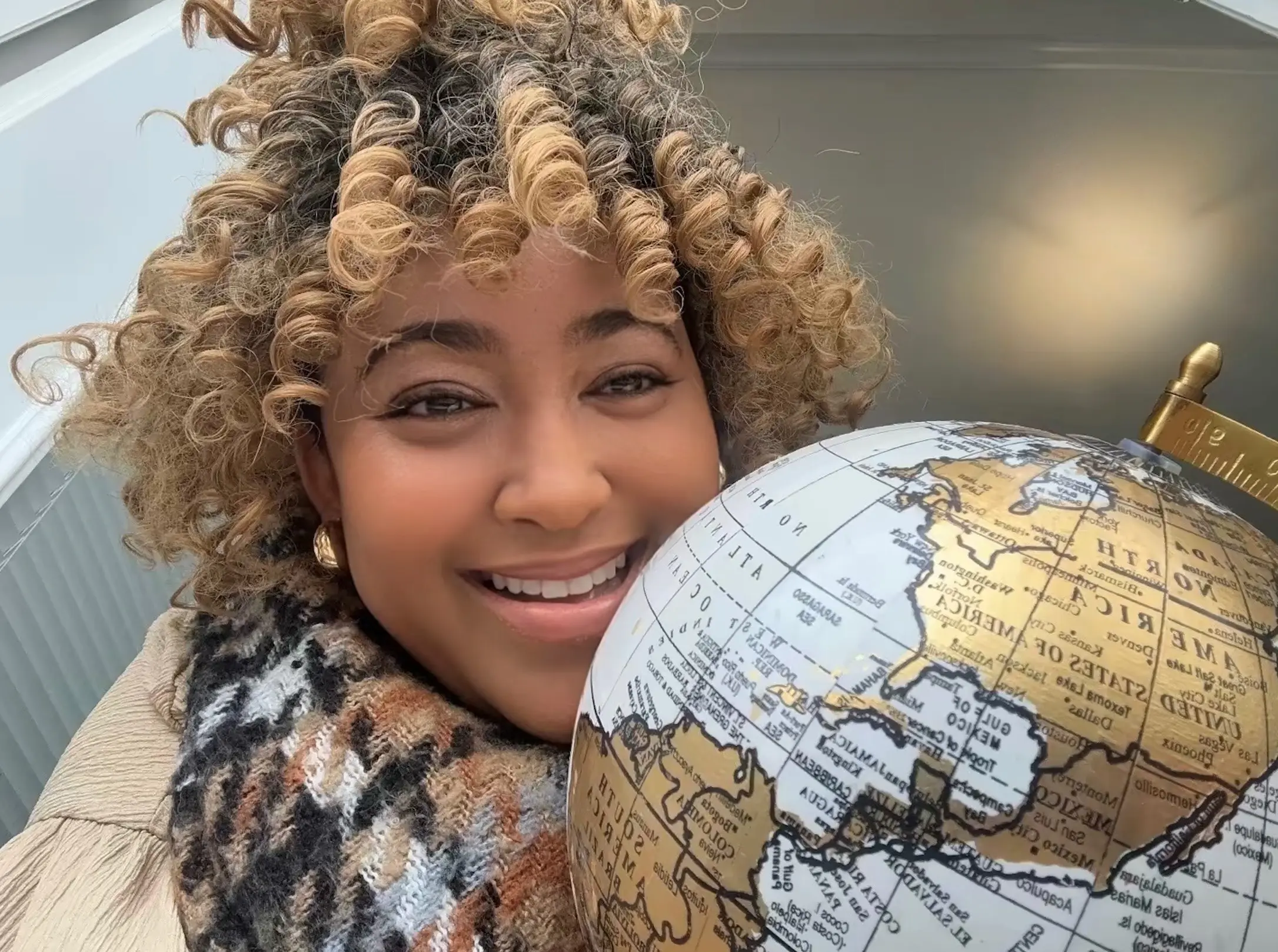 Smiling woman with blonde curly hair holding a vintage gold desktop globe indoors.