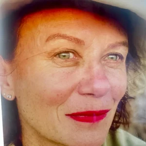 Close-up portrait of a smiling woman with green eyes, red lipstick, and a sun hat.