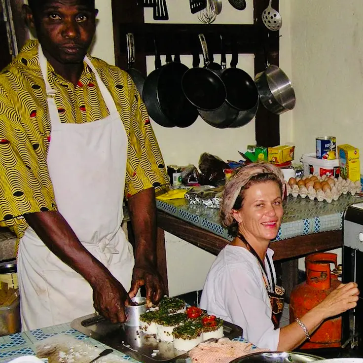 A smiling woman and a male chef in an apron preparing decorated, layered savory dishes on a tray in a kitchen with hanging pots and pans.
