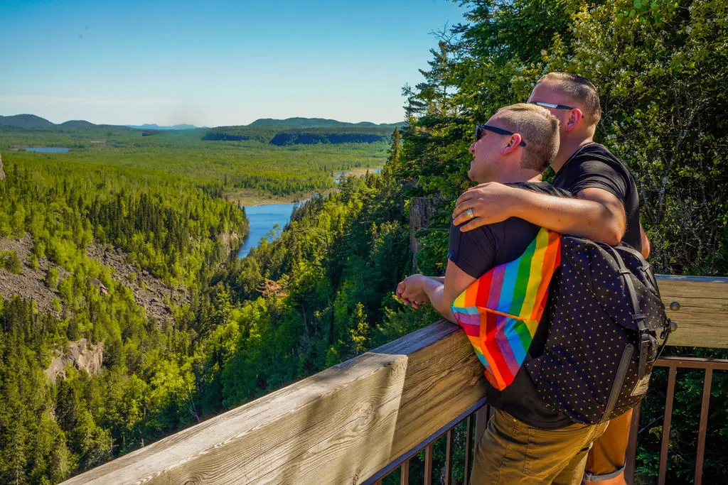 A couple with a Pride flag bandana embracing at a wooden scenic overlook viewing a lush green forest valley and river.