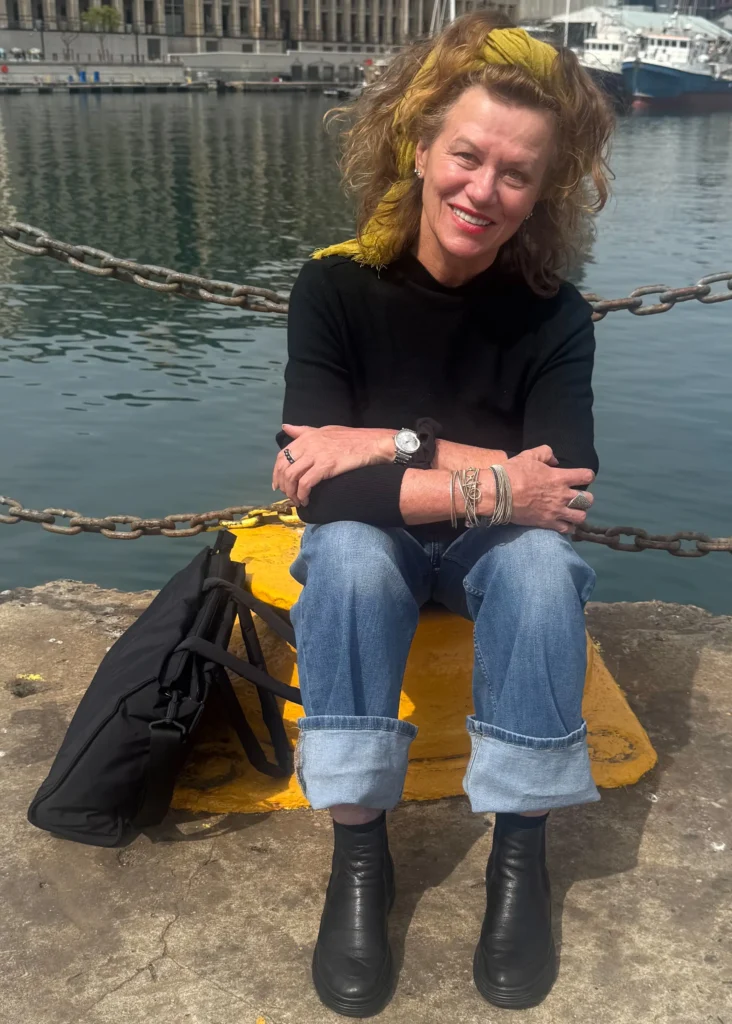 A smiling woman wearing a yellow headband, black sweater, and blue jeans sitting on a yellow harbor bollard by the water at a harbor.
