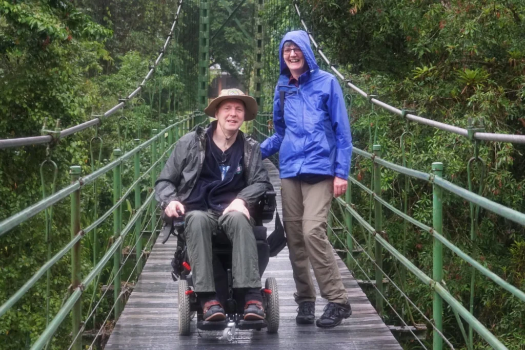 A man in a motorized power wheelchair wearing a sun hat and rain jacket alongside a woman in a blue raincoat smiling on an accessible wooden suspension bridge.