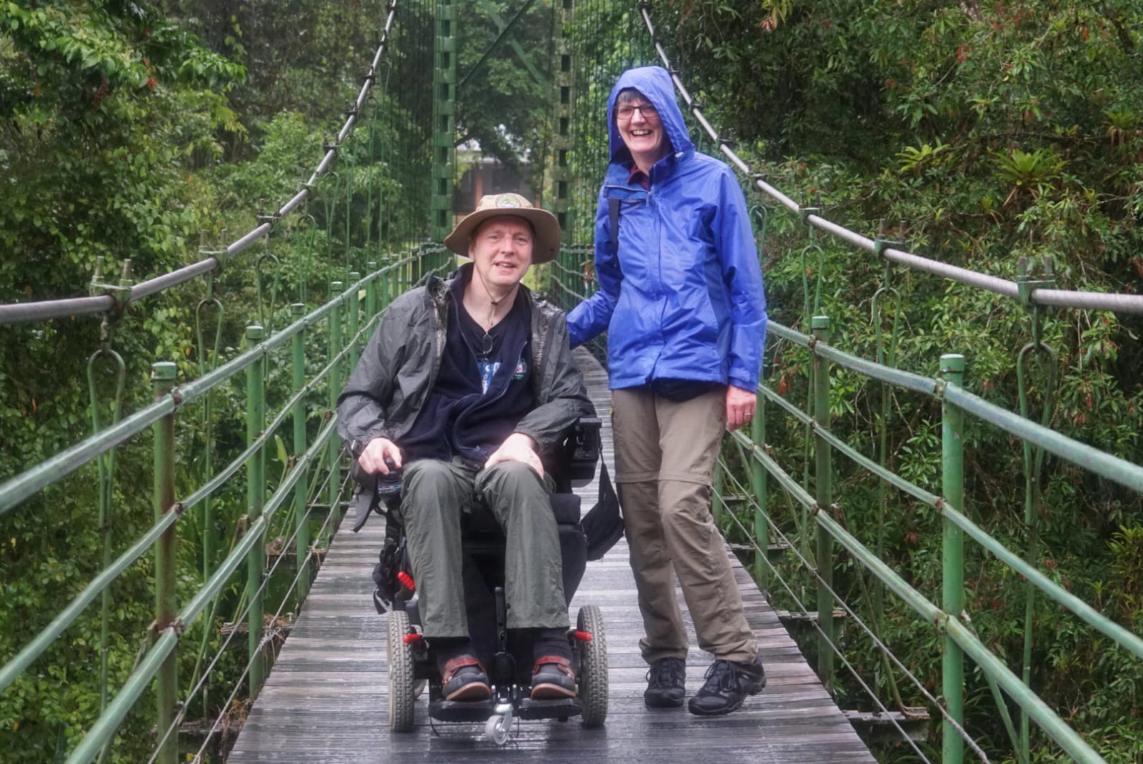A man in a motorized power wheelchair wearing a sun hat and rain jacket alongside a woman in a blue raincoat smiling on an accessible wooden suspension bridge.