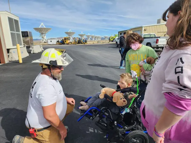 A firefighter kneeling down to talk with a young boy in a wheelchair at an outdoor site with satellite dishes in the background.