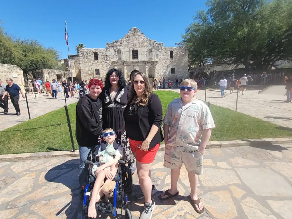 Family posing for a picture in front of the historic Alamo mission in San Antonio, Texas, with a child using a wheelchair in the foreground.
