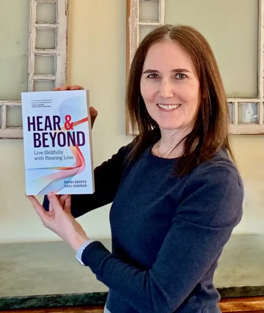 Author Shari Eberts smiling while holding up a copy of her book Hear and Beyond: Live Skillfully with Hearing Loss.