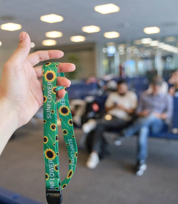 Hand holding a green Sunflower hidden disabilities lanyard in a blurred airport terminal setting.