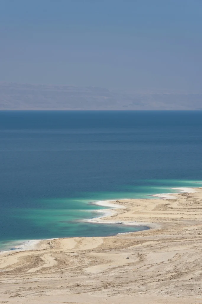 Scenic high-angle view of the Dead Sea coastline featuring turquoise water, white salt crust formations along the shoreline, and desert mountains under a clear blue sky.