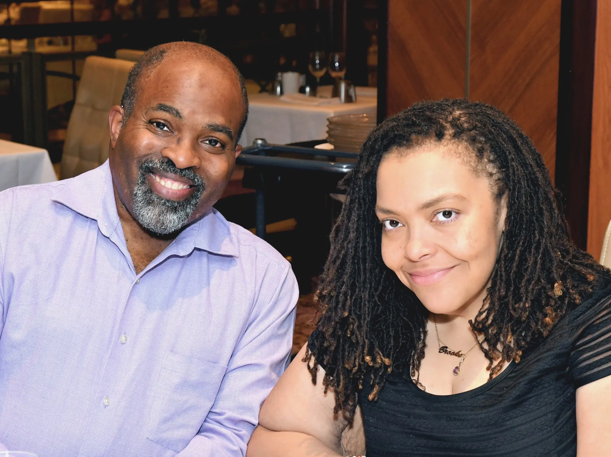 A close-up portrait of Aadam and Brooke smiling during a dinner date. Aadam is wearing a light purple button-down shirt, and Brooke is wearing a black top with a personalized name necklace.