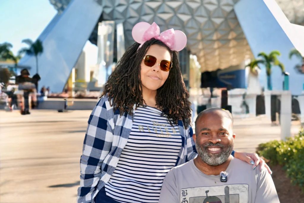 Brooke, wearing pink Minnie Mouse ears and a flannel shirt, stands smiling with her hand on the shoulder of her husband Aadam, a wheelchair user, in front of Epcot's Spaceship Earth.