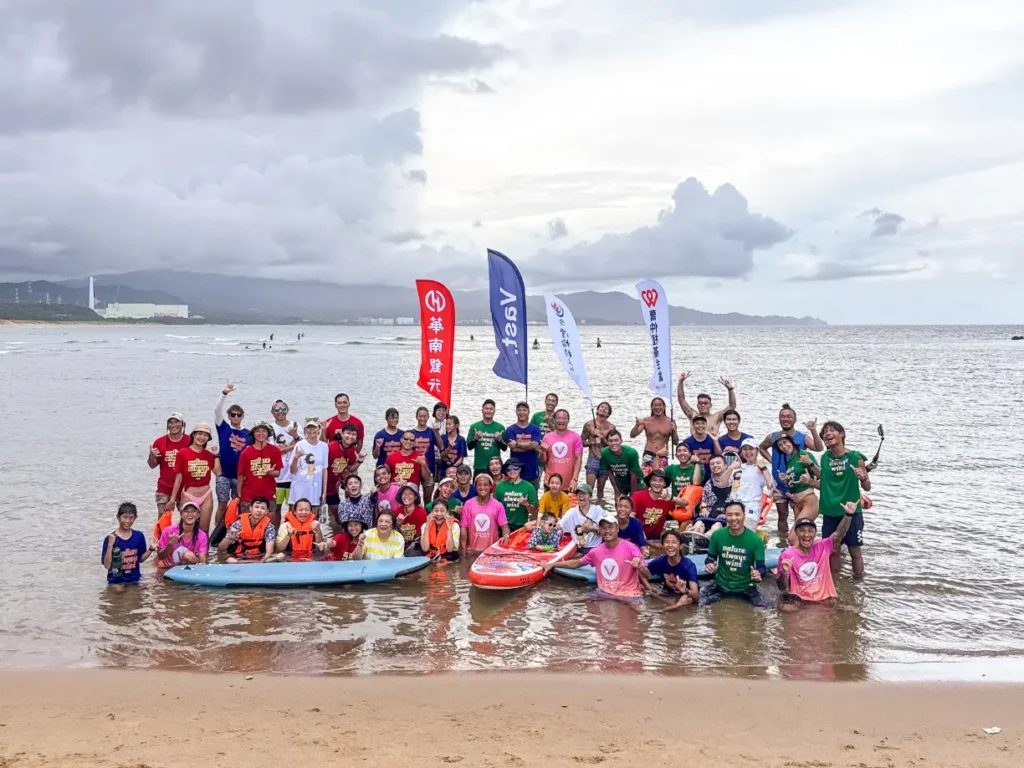 A large group of adaptive surfers, volunteers, and supporters posing together in shallow ocean water with colorful flags at a beach event.