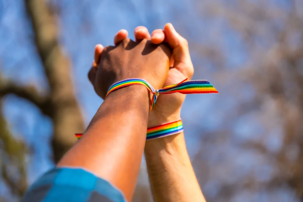 Two people holding hands raised high against the sky, both wearing colorful rainbow LGBTQ+ pride ribbon wristbands.