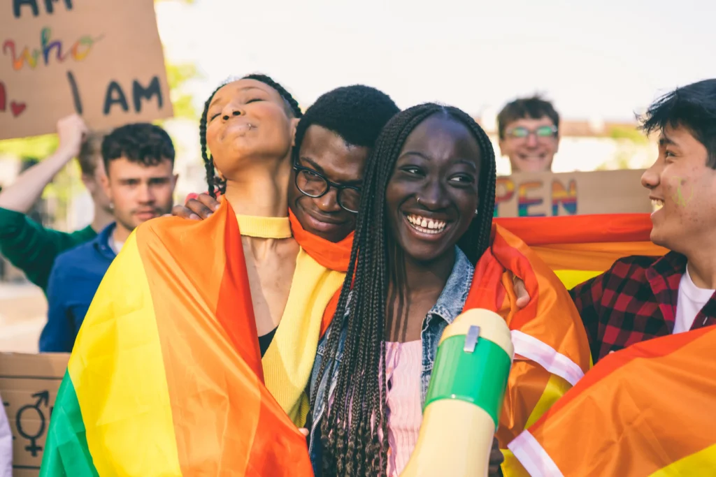 Group of diverse young adults smiling and hugging wrapped in a rainbow LGBTQ+ pride flag at an outdoor rally.