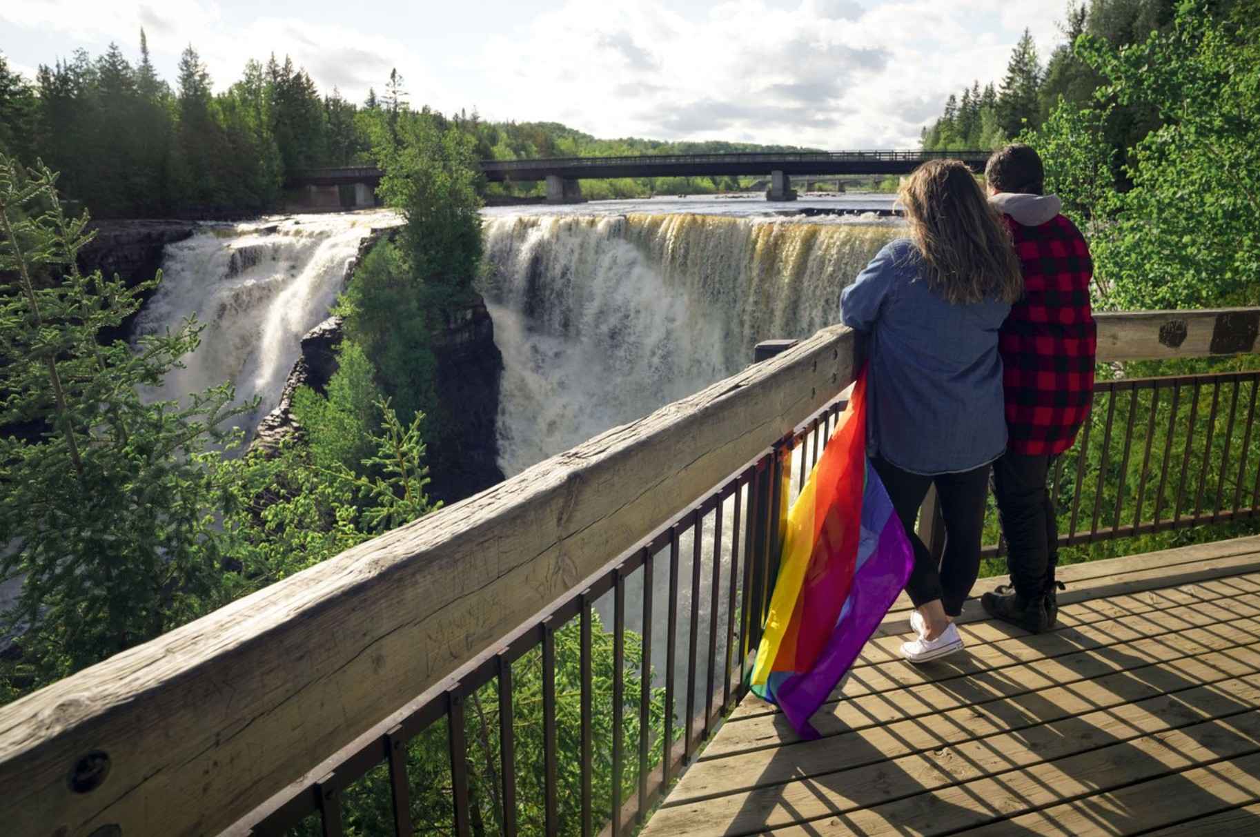 Two people stand together at a wooden lookout overlooking a large waterfall surrounded by lush forest, with a Pride flag draped over the railing beside them.
