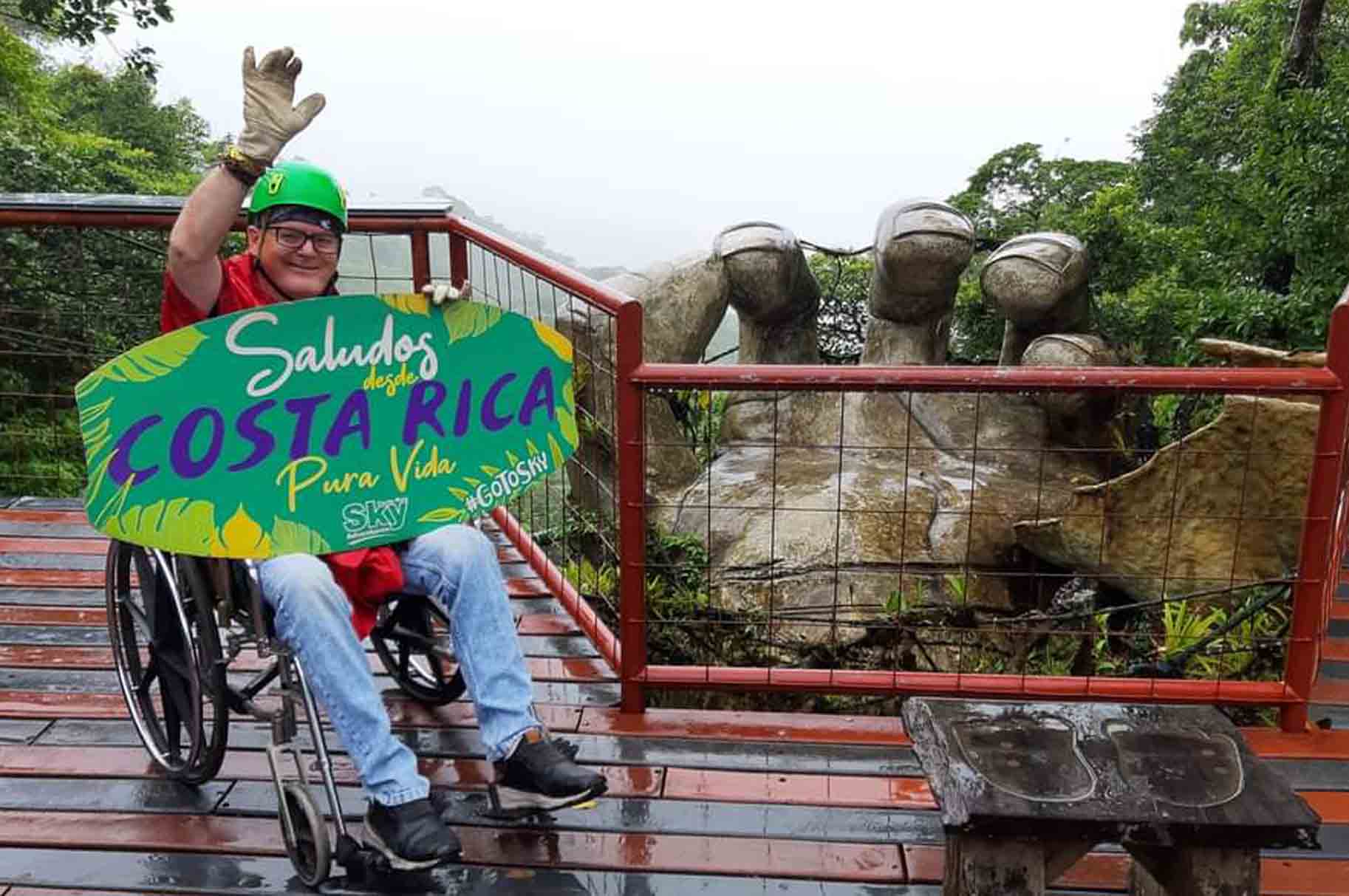 Wheelchair user holding a colourful “Saludos desde Costa Rica” sign on an accessible viewing platform surrounded by lush rainforest.