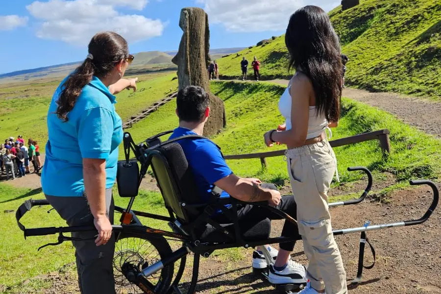 A man seated in a specialized all-terrain wheelchair with long carrying handles looks toward a large moai statue on a grassy hillside, flanked by a woman guide in a teal shirt pointing toward the statue and another woman with long dark hair standing beside him. Other visitors are visible walking on a path in the background under a partly cloudy sky.