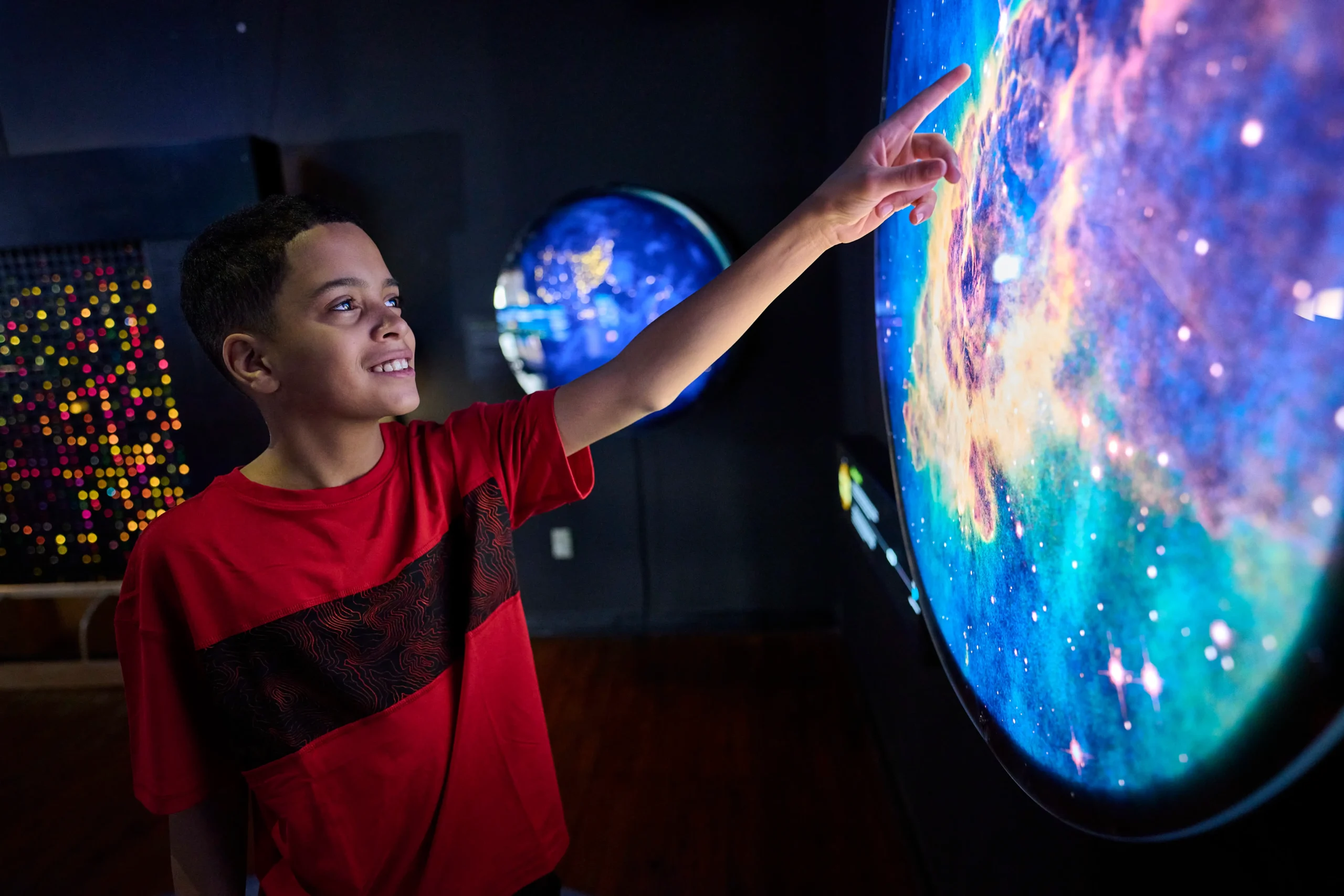 A young boy smiling and pointing at a bright, colorful interactive space exhibit on a wall.