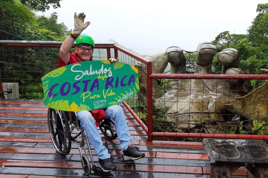 A man in a wheelchair wearing a green zip-line helmet waves cheerfully while holding a 'Saludos desde Costa Rica' sign on a wooden platform, with large hand-shaped sculptures visible in the misty forest behind him.